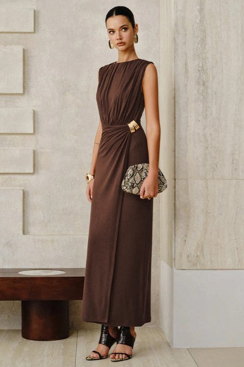 Woman in a brown dress holding a clutch against a neutral wall.