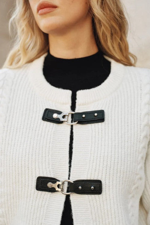 Close-up of a person wearing a white textured cardigan with black leather fastenings.