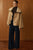 Woman wearing a beige coat over a brown turtleneck and dark blue pants against a wooden paneled wall.