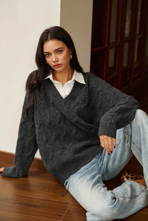 Woman wearing a dark gray sweater over a white shirt and light blue jeans, sitting on a wooden floor.